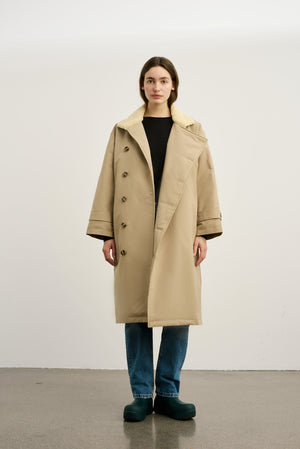 A model wears a beige MEOTINE coat with a soft collar, paired with blue jeans and green clogs, set against a plain background.