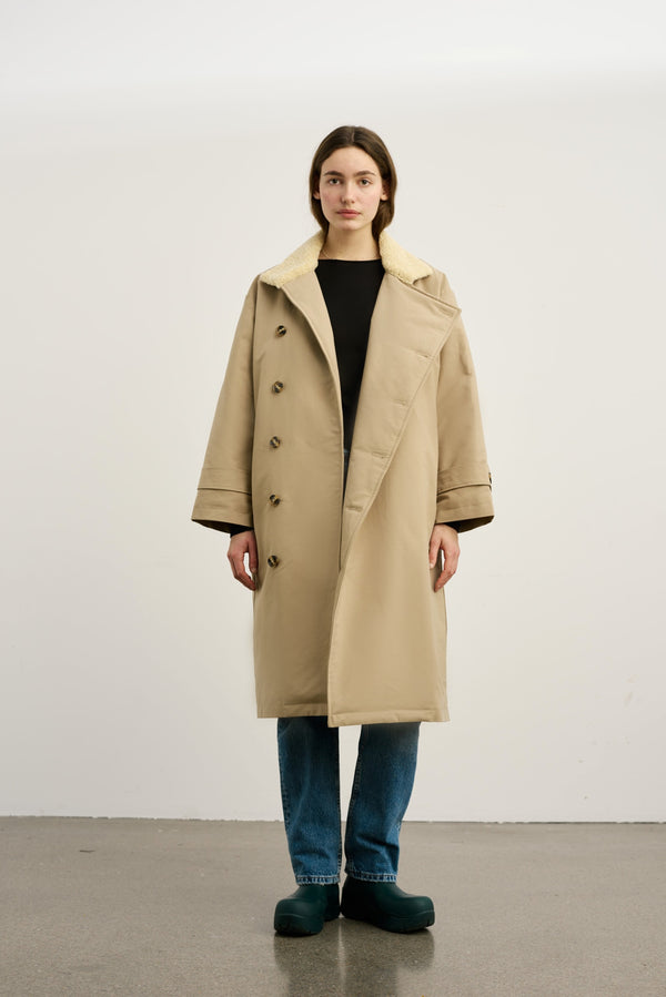 A model wears a beige MEOTINE coat with a soft collar, paired with blue jeans and green clogs, set against a plain background.