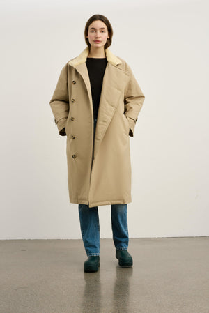 A model wearing a beige MEOTINE coat with a soft collar, paired with a black top and blue jeans, standing against a neutral background.