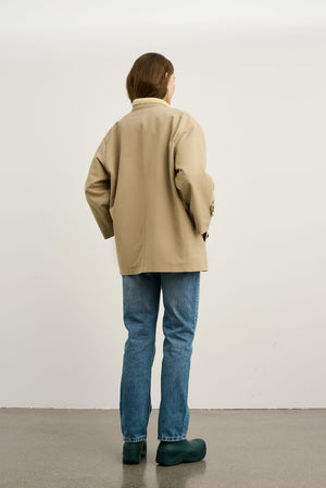 A woman in a beige oversized jacket and blue jeans stands with her back to the camera, showcasing a casual MEOTINE style.