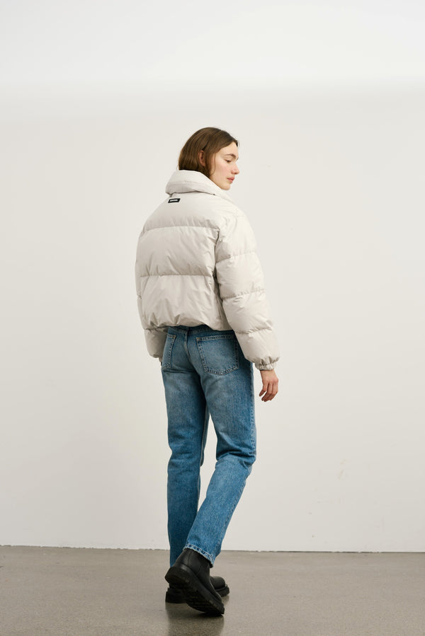 A model wearing a light gray puffer jacket with a high collar, paired with blue jeans and black boots, showcasing MEOTINE style.