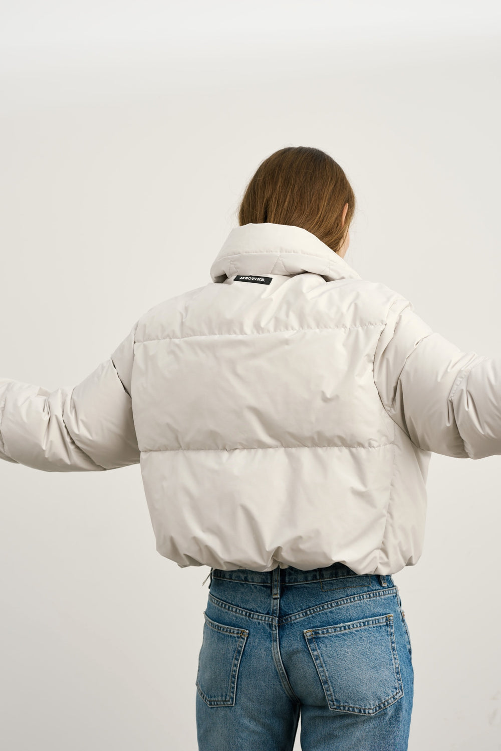 A model wears a light beige puffer jacket paired with blue jeans and black shoes, showcasing a stylish winter look by MEOTINE.