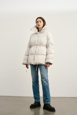 A model wears a light beige puffer jacket with a cinched waist and high collar, paired with blue jeans and black boots. MEOTINE.