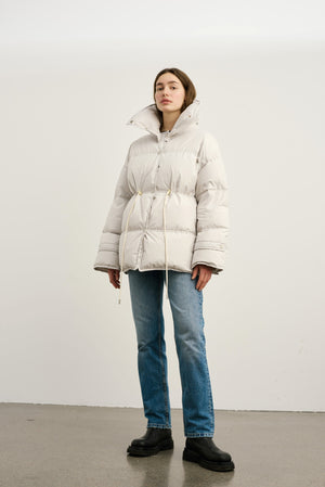 A model wears a stylish, oversized light gray puffer jacket with a cinched waist, paired with blue jeans. MEOTINE brand.