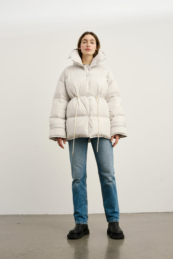 A model wears a stylish, oversized white puffer jacket with a cinched waist, paired with blue jeans and black boots by MEOTINE.