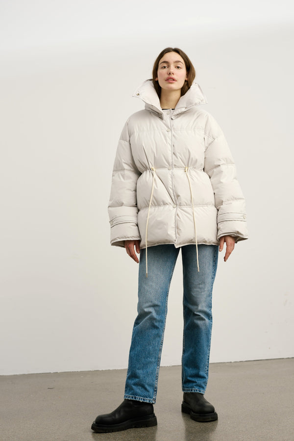 A model wears a stylish, oversized light gray puffer jacket with a cinched waist, paired with blue jeans and black boots. MEOTINE.