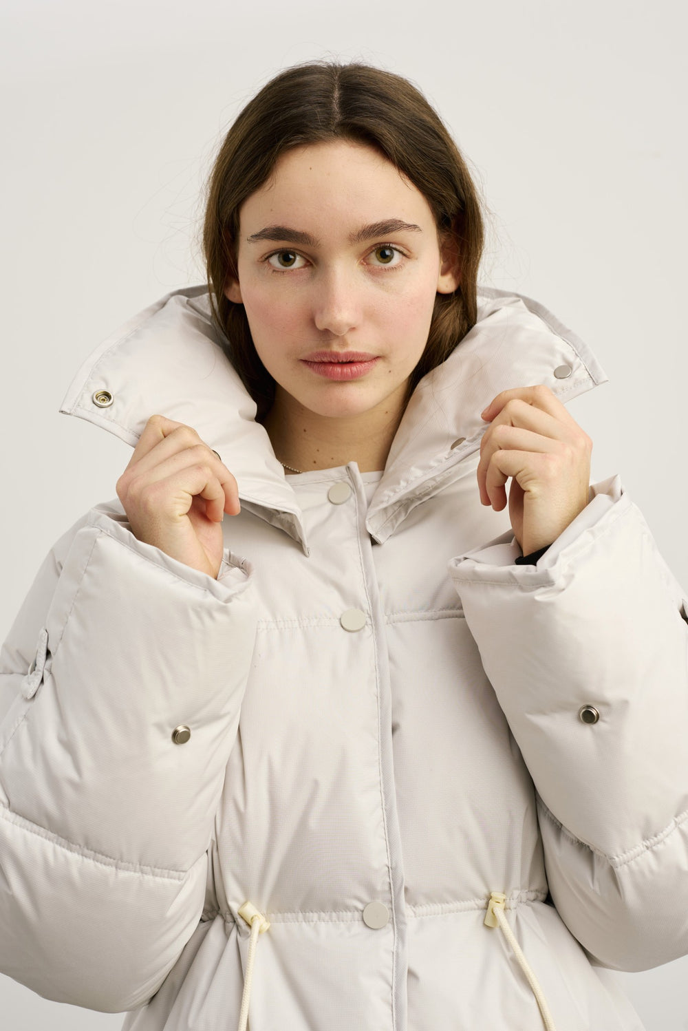A model wears a stylish, oversized light gray puffer jacket with a cinched waist, paired with blue jeans and black boots. MEOTINE.