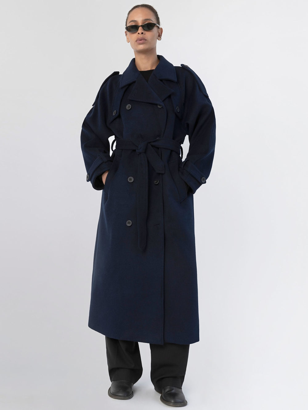 A model wears a stylish navy trench coat with a belt, paired with black pants and sunglasses, showcasing MEOTINE's chic design.