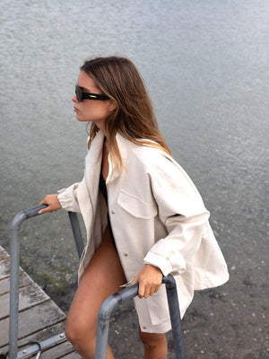 A woman in a beige oversized shirt and sunglasses steps onto a dock by the water, showcasing a relaxed summer vibe with MEOTINE style.