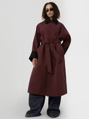 A model showcases a stylish burgundy coat with a tie waist and contrasting black cuffs, paired with wide-leg denim pants. MEOTINE.
