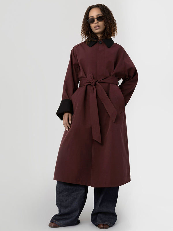 A model showcases a stylish burgundy coat with a tie waist and contrasting black cuffs, paired with wide-leg denim pants. MEOTINE.