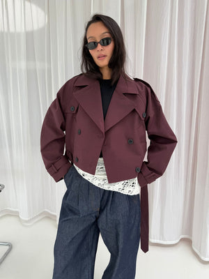 Chic model wearing a burgundy cropped trench coat over a white lace top, paired with wide-leg denim pants and sunglasses. MEOTINE.