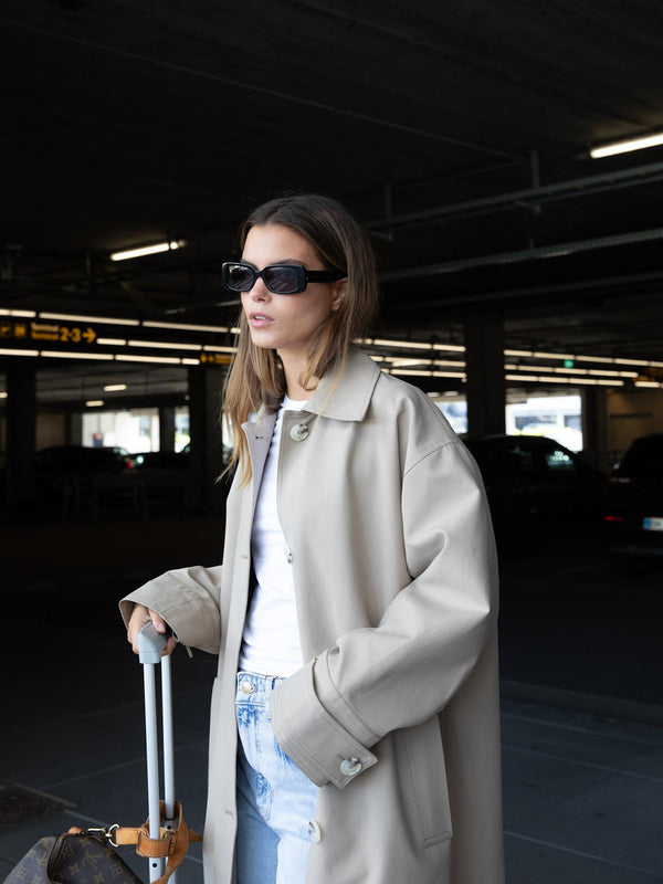 A stylish woman in a beige oversized coat and sunglasses stands with a suitcase, embodying modern travel chic by MEOTINE.
