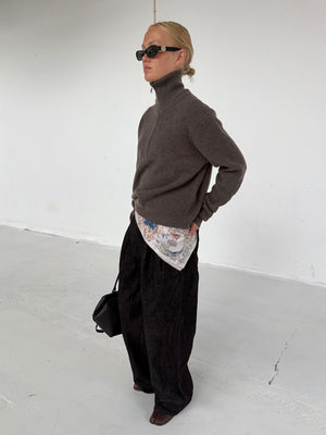 A model showcases a stylish outfit featuring a brown zip-up sweater, patterned shirt, and wide black pants, complemented by sunglasses.
