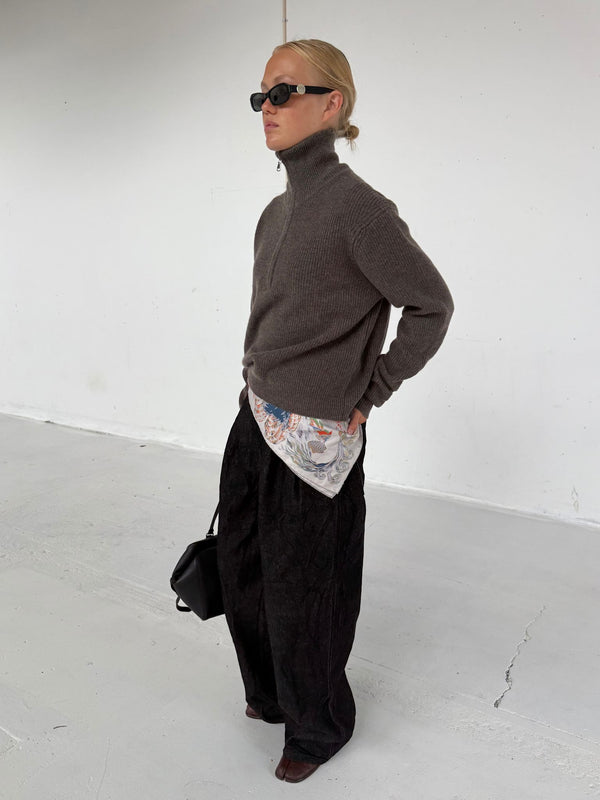 A model showcases a stylish outfit featuring a brown zip-up sweater, patterned shirt, and wide black pants, complemented by sunglasses.