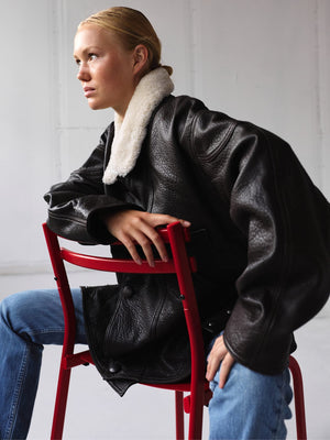 A model wearing a stylish black leather jacket with a cozy shearling collar, seated on a red chair, showcasing MEOTINE's chic design.
