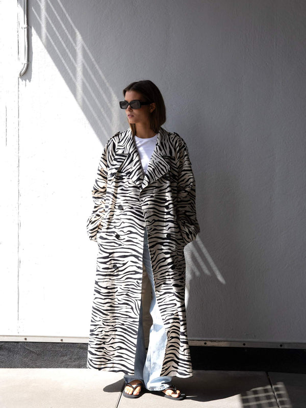 A model wearing a stylish zebra print coat by MEOTINE, paired with light jeans and sunglasses, stands against a white wall.