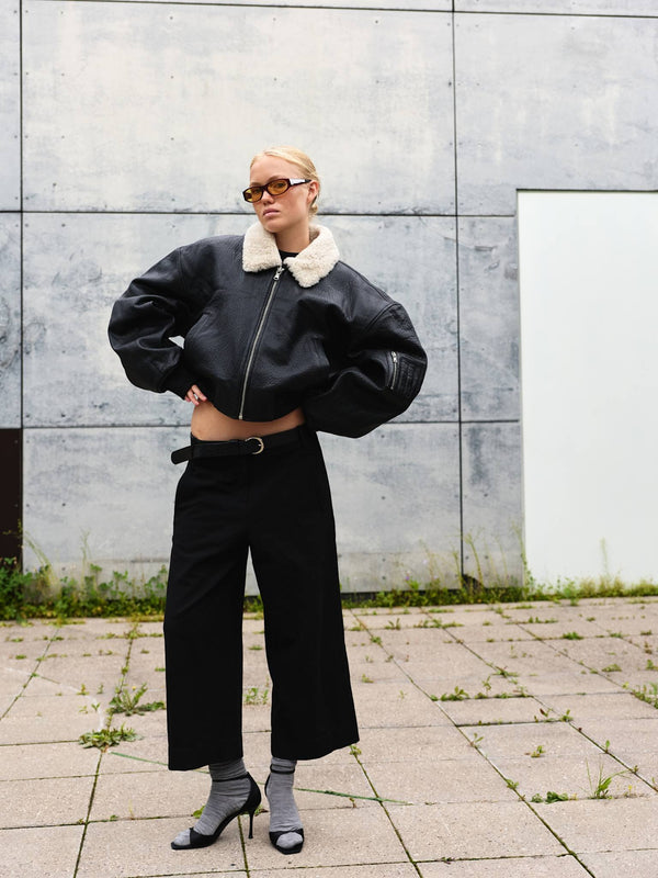 A model showcases a stylish black leather bomber jacket with a fur collar, paired with wide-leg black trousers and chic heels.