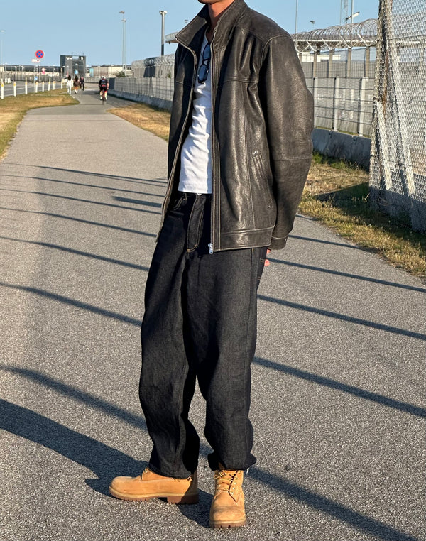 A person stands on a path wearing a black leather jacket, white t-shirt, loose dark pants, and tan boots, showcasing MEOTINE style.