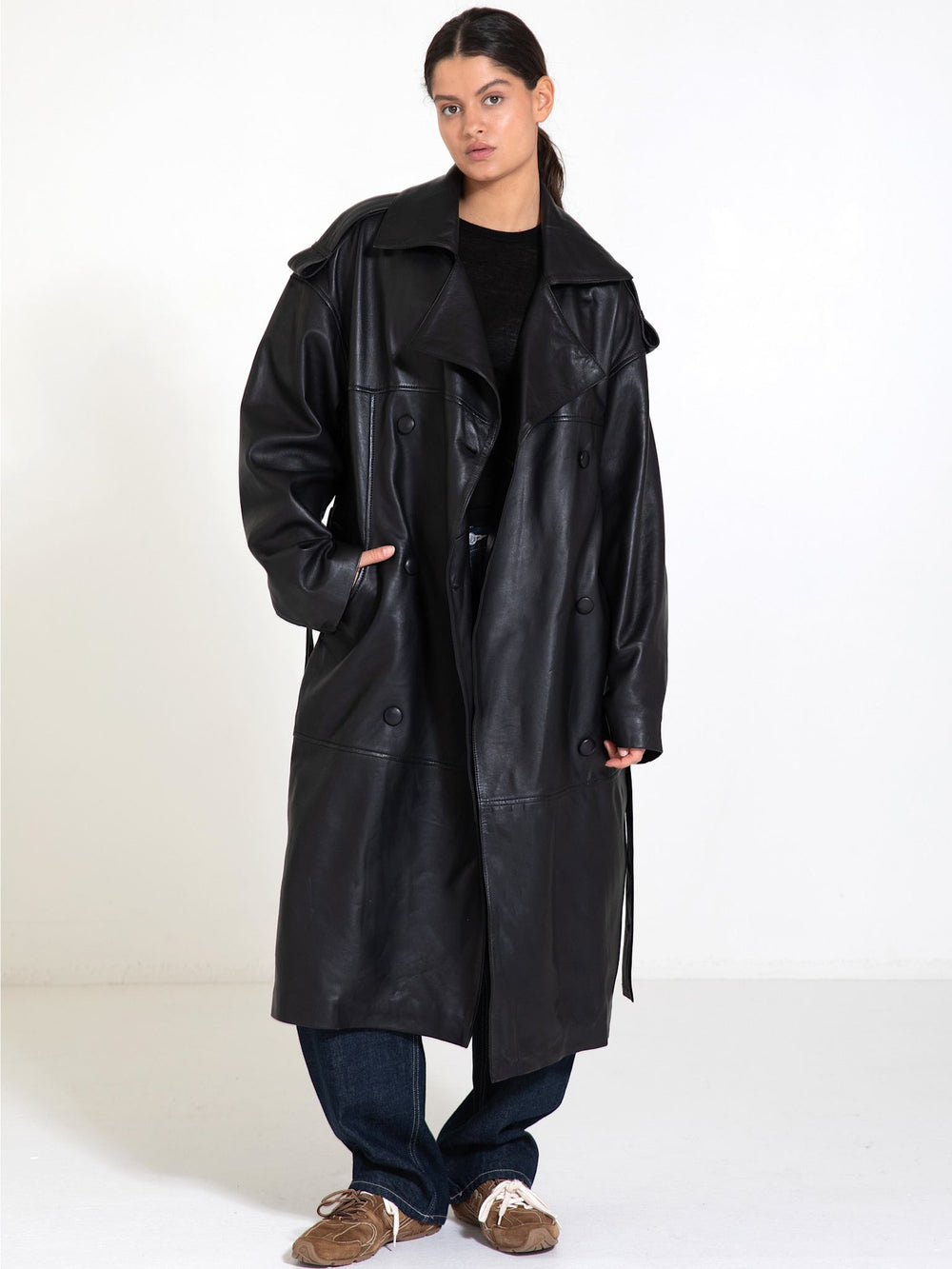 A model showcases a stylish black leather trench coat by MEOTINE, paired with casual jeans and sneakers against a neutral backdrop.