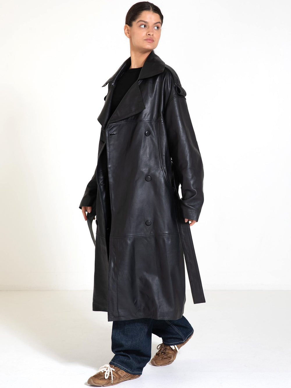 A model showcases a stylish black leather trench coat by MEOTINE, paired with casual jeans and sneakers against a neutral backdrop.