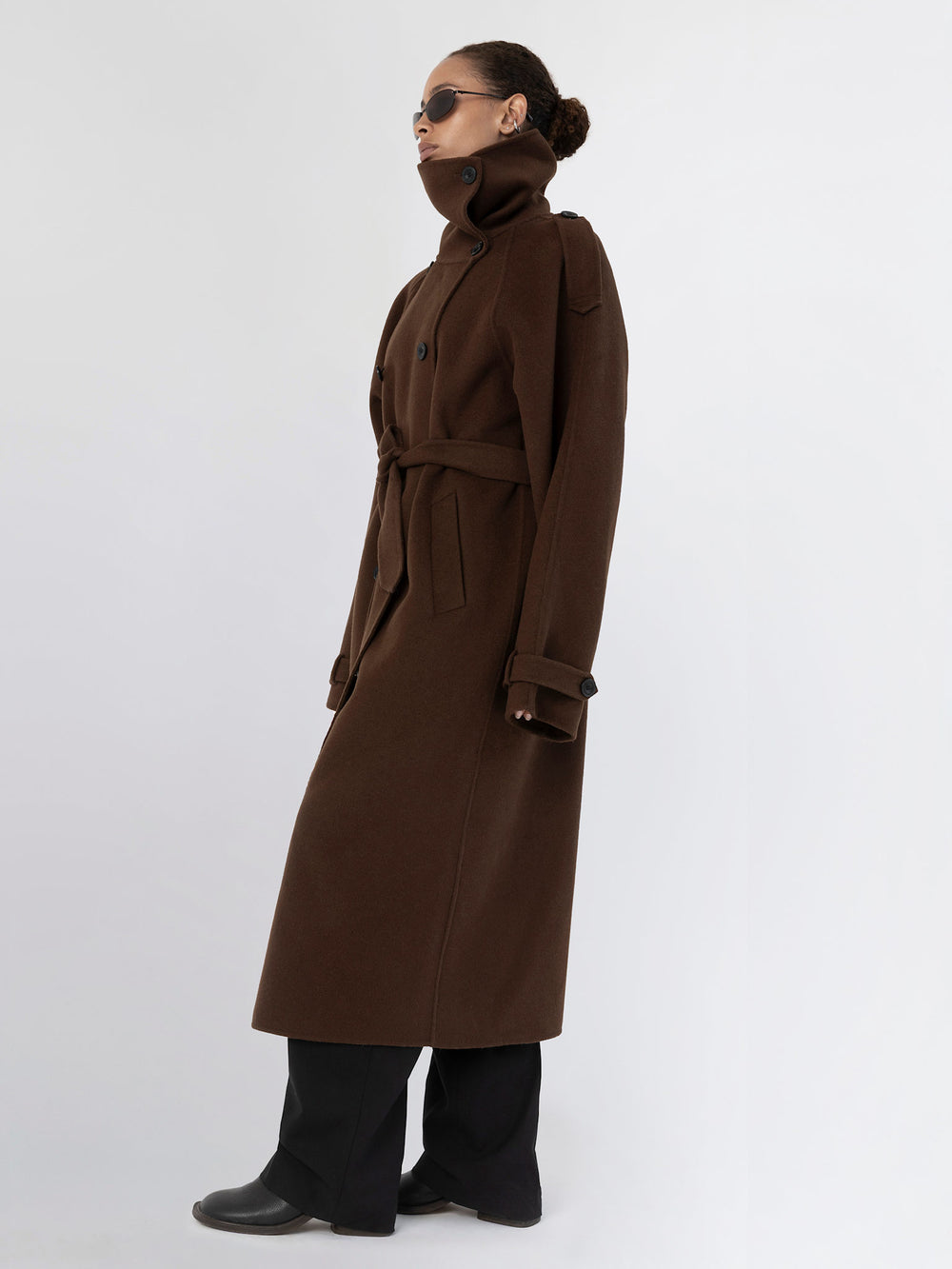 A model showcases a stylish brown trench coat with a high collar and belt, paired with black pants and sunglasses, by MEOTINE.