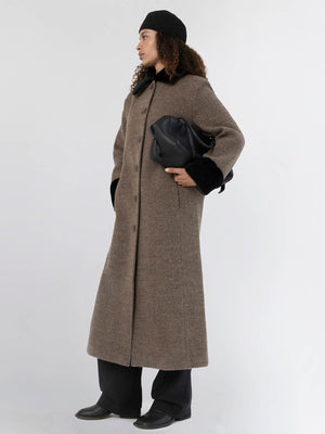 A model wearing a long brown coat with a black fur collar, paired with black pants and a stylish bag, showcasing MEOTINE's elegance.