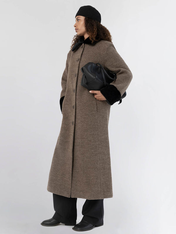 A model wearing a long brown coat with a black fur collar, paired with black pants and a stylish bag, showcasing MEOTINE's elegance.