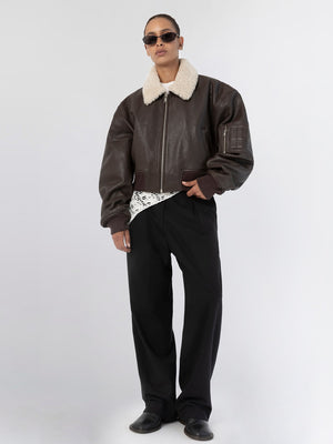 A model wearing a brown leather bomber jacket with a shearling collar, paired with black pants and sunglasses, showcasing MEOTINE style.