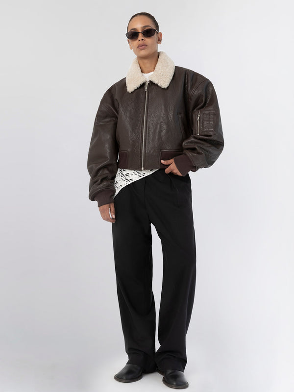 A model wearing a brown leather bomber jacket with a shearling collar, paired with black pants and sunglasses, showcasing MEOTINE style.