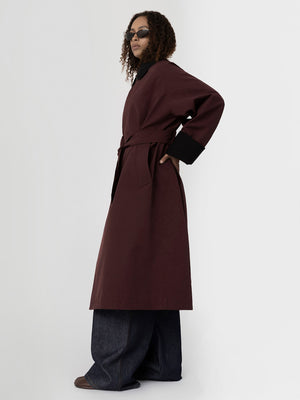 A model poses in a burgundy MEOTINE coat with a belted waist, paired with wide-leg denim pants and stylish sunglasses.