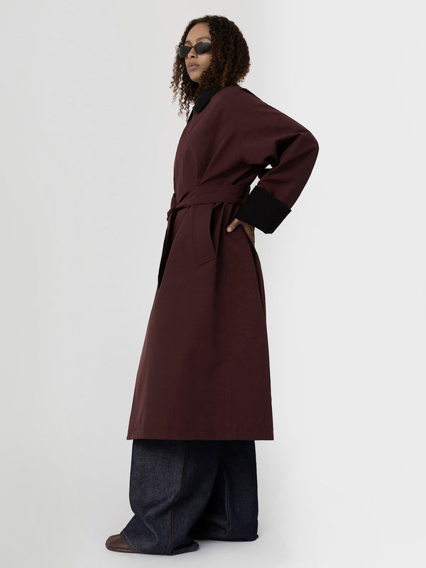 A model poses in a burgundy MEOTINE coat with a belted waist, paired with wide-leg denim pants and stylish sunglasses.
