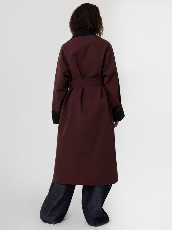 A model showcases a stylish burgundy trench coat with a black collar and belt, paired with wide-leg denim pants. MEOTINE.