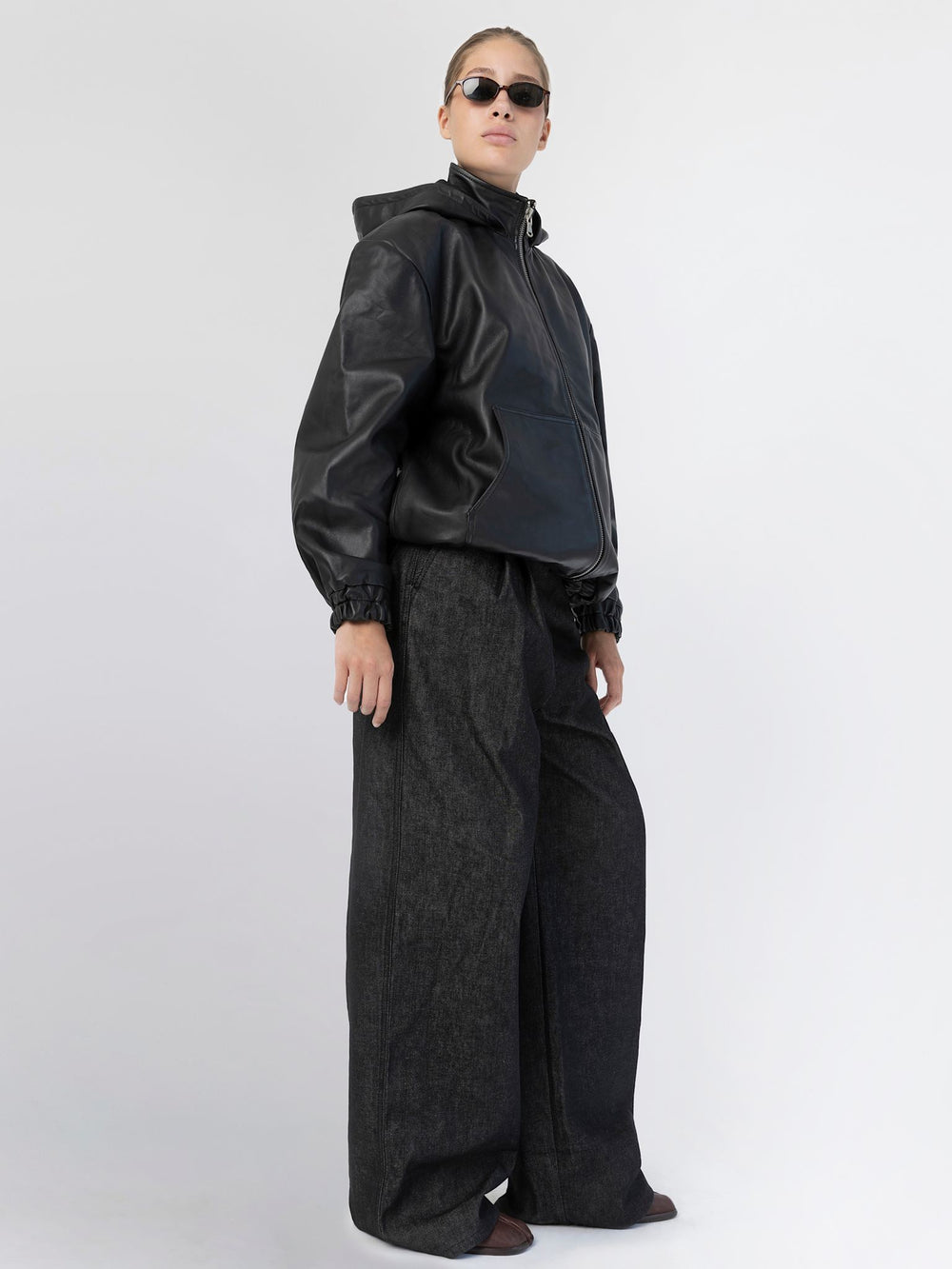 Stylish black leather jacket with a hood, featuring a zip closure and relaxed fit, paired with sleek sunglasses. MEOTINE.