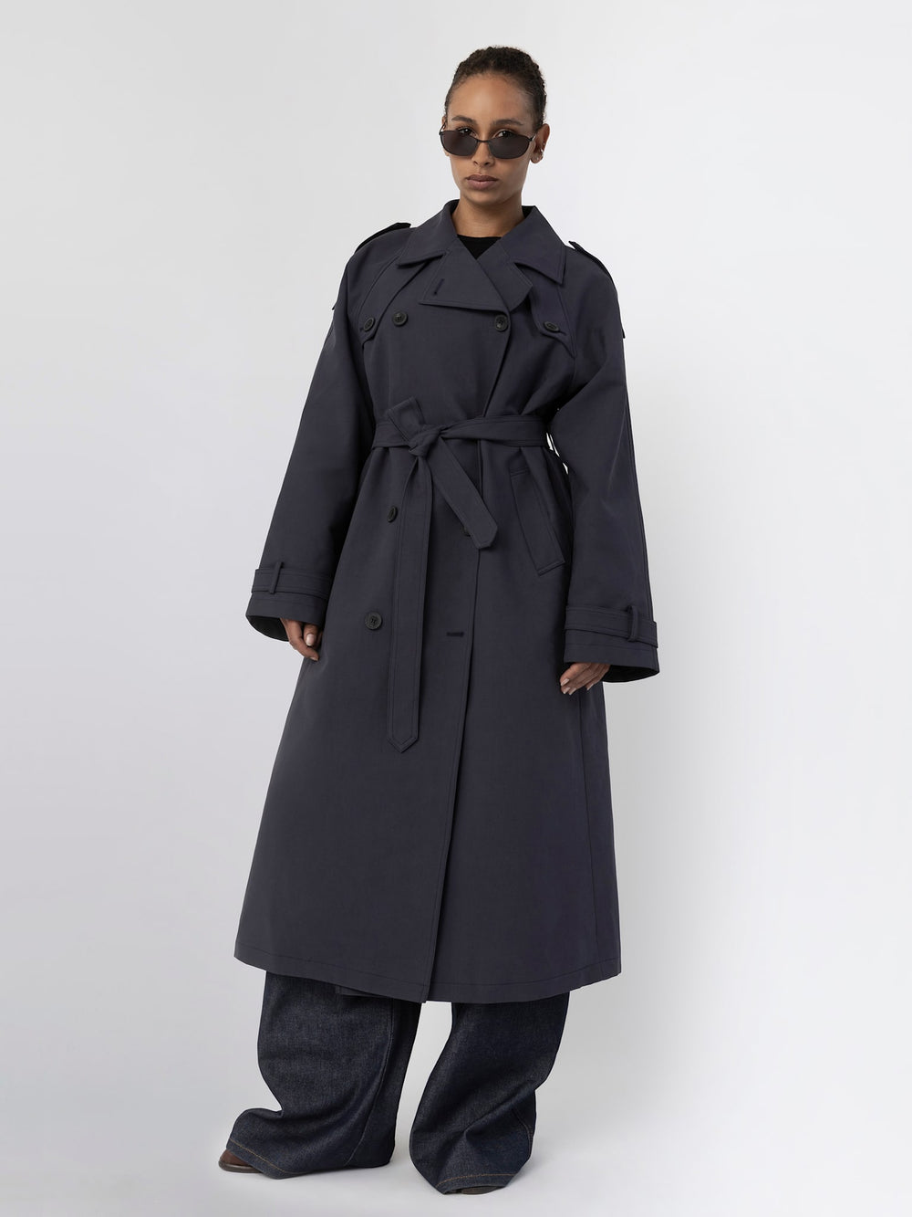 A model showcases a stylish dark trench coat with a belt, paired with wide-leg jeans and sunglasses, representing MEOTINE's chic aesthetic.