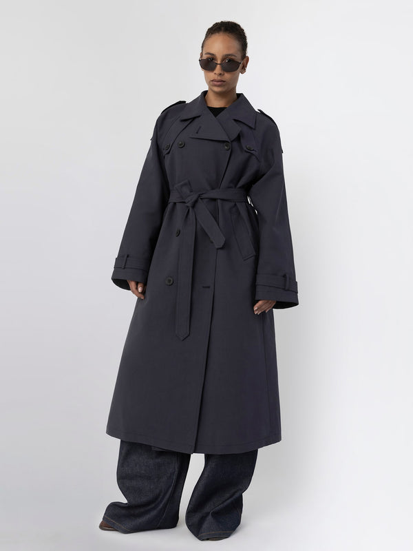 A model showcases a stylish dark trench coat with a belt, paired with wide-leg jeans and sunglasses, representing MEOTINE's chic aesthetic.