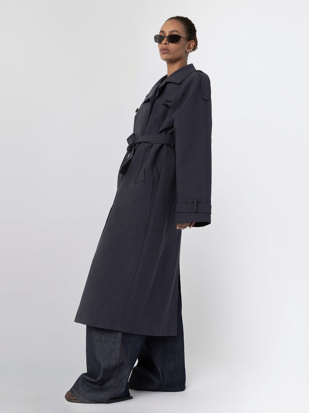 A model showcases a stylish dark trench coat with a belt, paired with wide-leg jeans and sunglasses, representing MEOTINE's chic aesthetic.