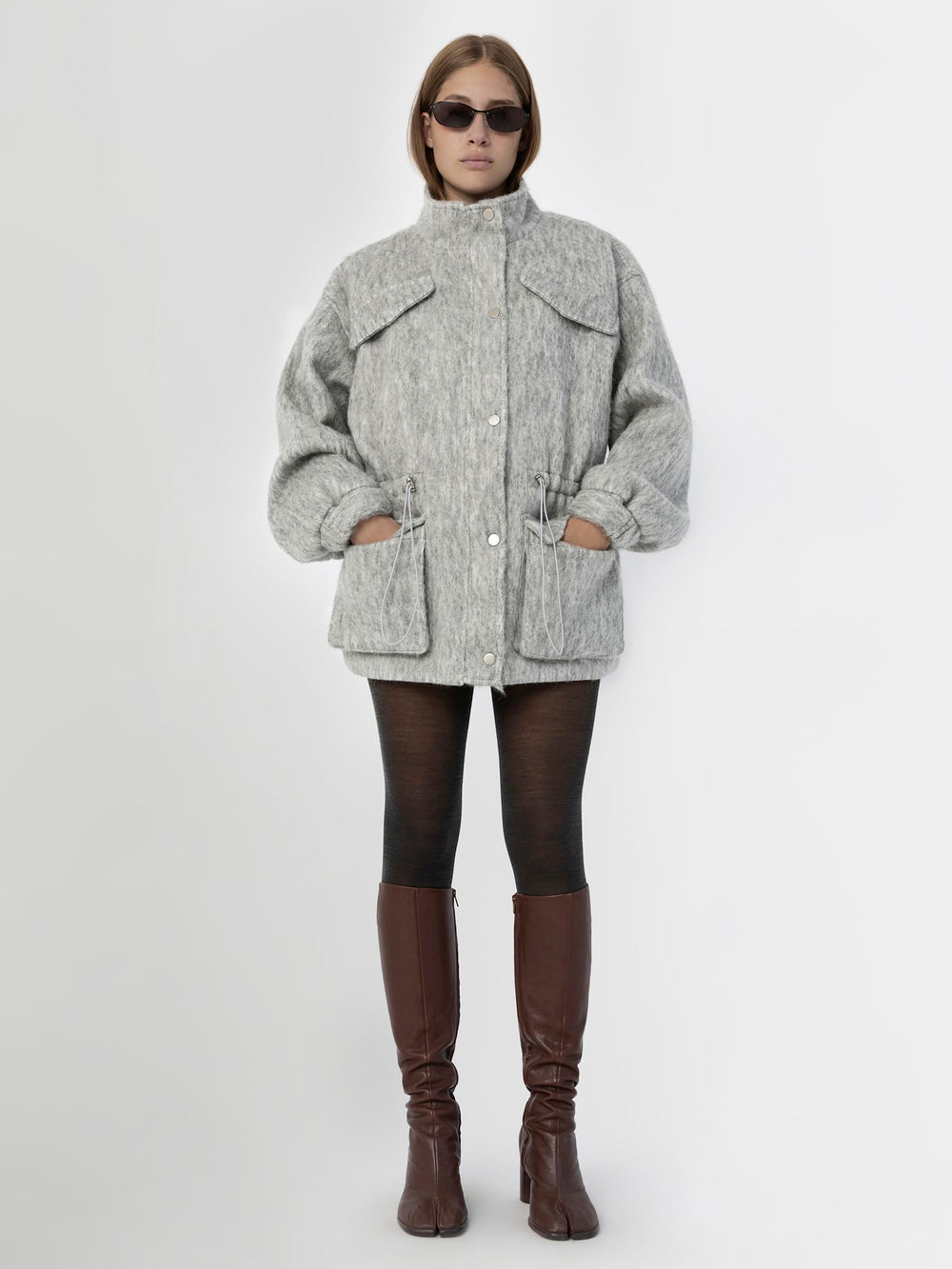 A model showcases a stylish gray MEOTINE jacket with a high collar, front pockets, and a cinched waist, paired with blue jeans.