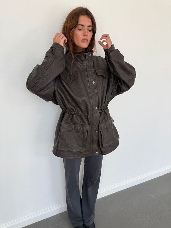 A model wears a stylish brown MEOTINE jacket with a cinched waist and large pockets, paired with gray pants against a plain backdrop.