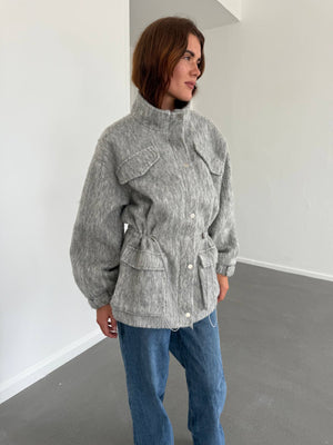 A model showcases a stylish gray MEOTINE jacket with a high collar, front pockets, and a cinched waist, paired with blue jeans.