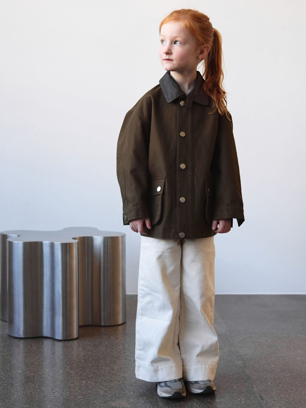 A child wearing a brown MEOTINE jacket with a contrasting collar, featuring large buttons and front pockets, stands against a neutral backdrop.
