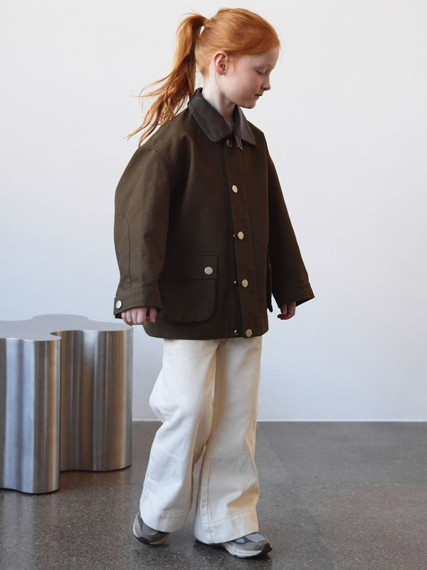 A child wearing a brown MEOTINE jacket with a gray collar, paired with white pants, walking in a modern indoor space.