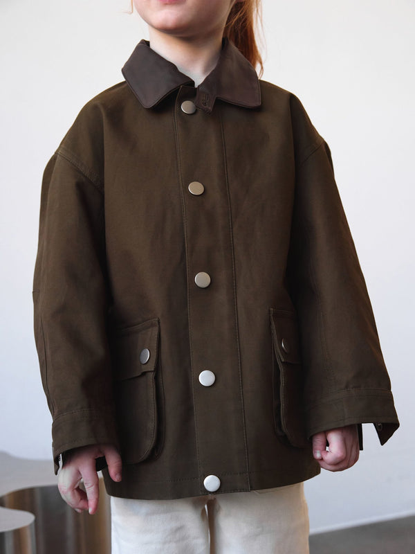 A child wearing a brown MEOTINE jacket with a contrasting collar, featuring large buttons and front pockets, stands against a neutral backdrop.