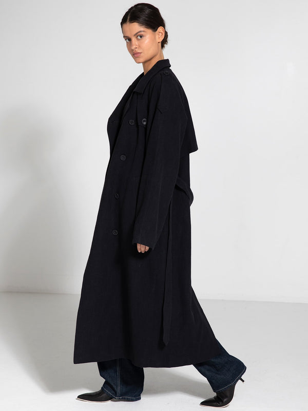 A model showcases a long, dark coat with a relaxed fit and oversized sleeves, styled with jeans and sleek shoes, by MEOTINE.