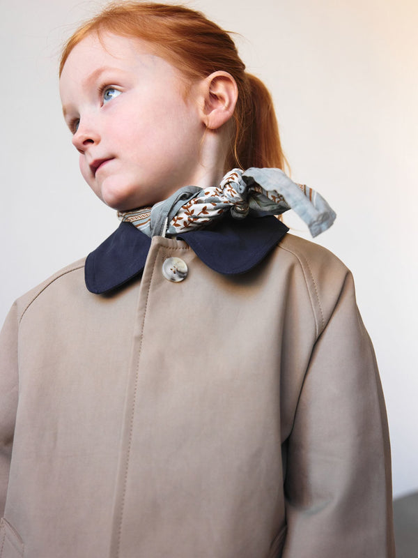 A young girl with red hair wears a beige coat with a navy collar, complemented by a patterned scarf, showcasing MEOTINE's stylish design.