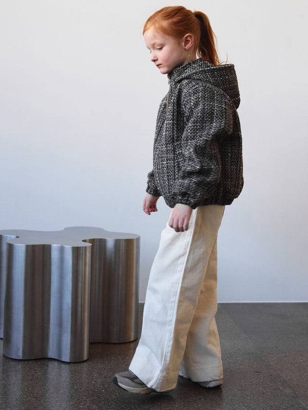 A girl in a textured black and white hoodie and white wide-leg pants stands beside a sculptural silver table, showcasing MEOTINE style.