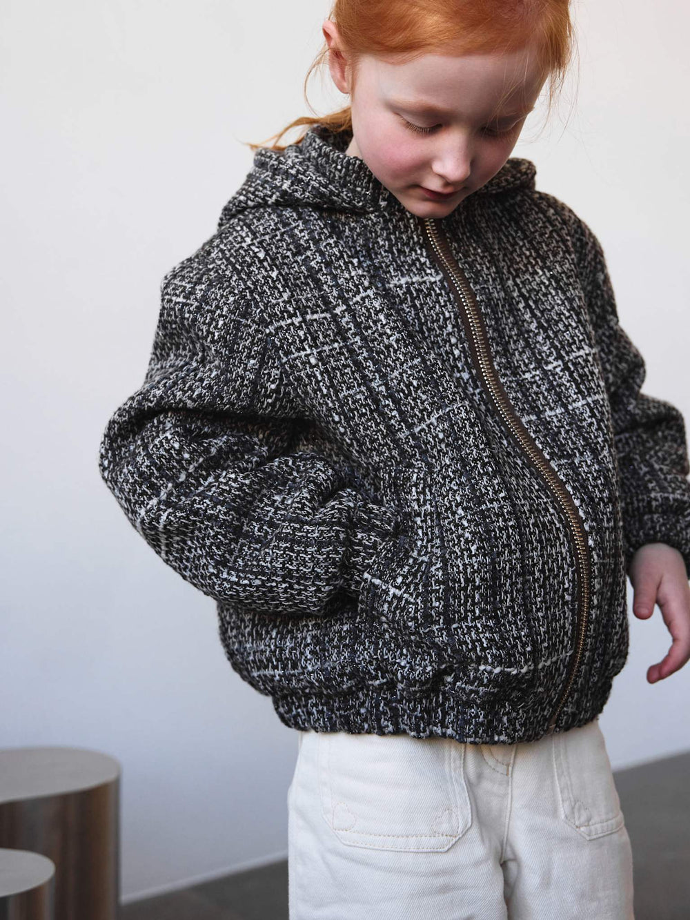 A girl in a textured black and white hoodie and white wide-leg pants stands beside a sculptural silver table, showcasing MEOTINE style.