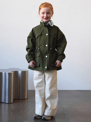 A child wearing a green MEOTINE jacket with pockets, paired with white pants, stands against a minimalist background.