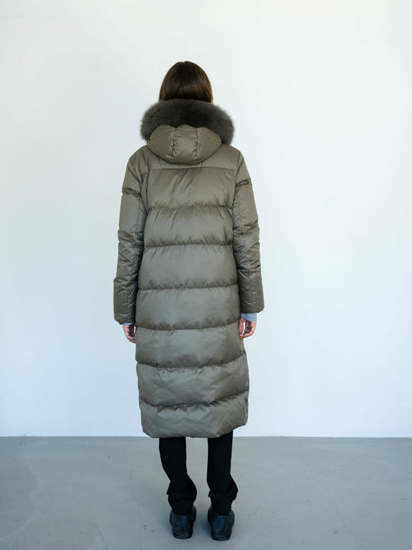 A woman stands with her back to the camera, wearing a long, olive green puffer coat with a fur-lined hood by MEOTINE.