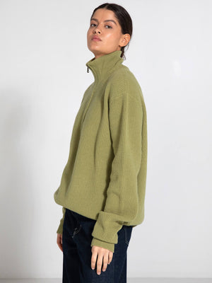 A model showcases a cozy, olive green MEOTINE sweater with a high collar, paired with dark jeans against a neutral background.
